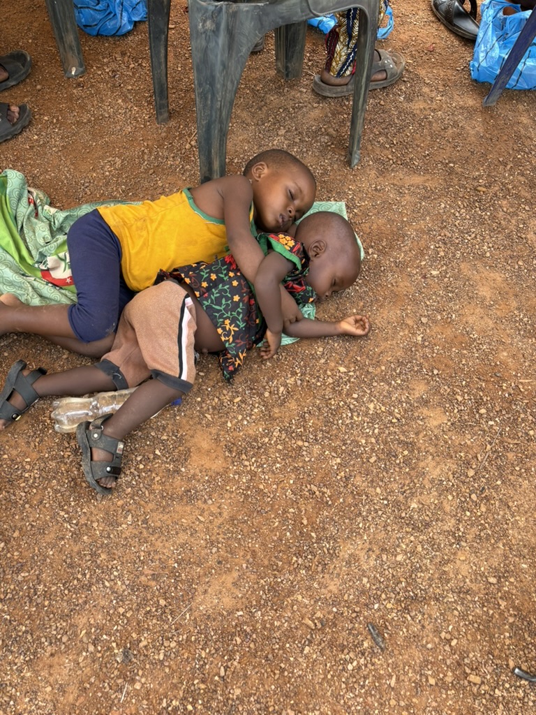 Two small children asleep on the red earth beneath plastic chairs at a medical mission.