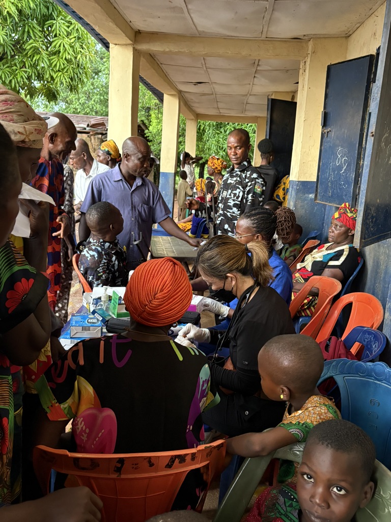 A medical mission in progress beneath a covered porch, mothers and children waiting to be seen.